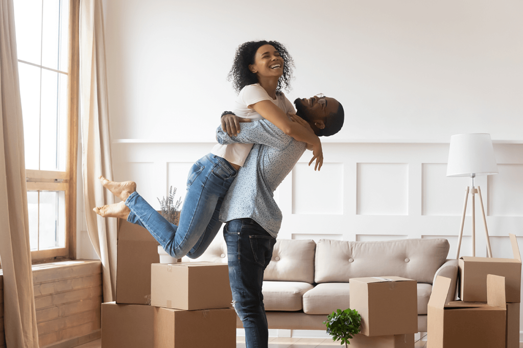 Young man hositing young woman off the ground in a newly bought house with unboxed boxes scattered
