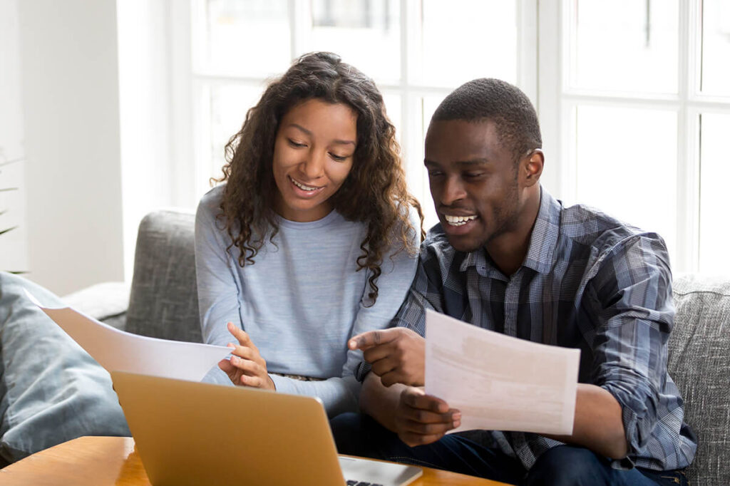 Young couple sitting on couch looking at laptop and holding financial documents